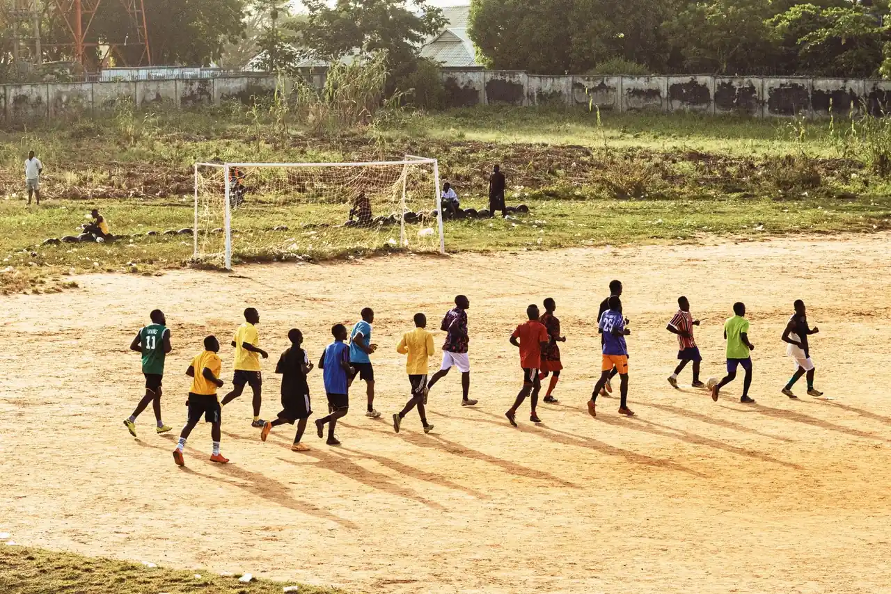 A group of young men practicing soccer on a dirt field under the sun.
